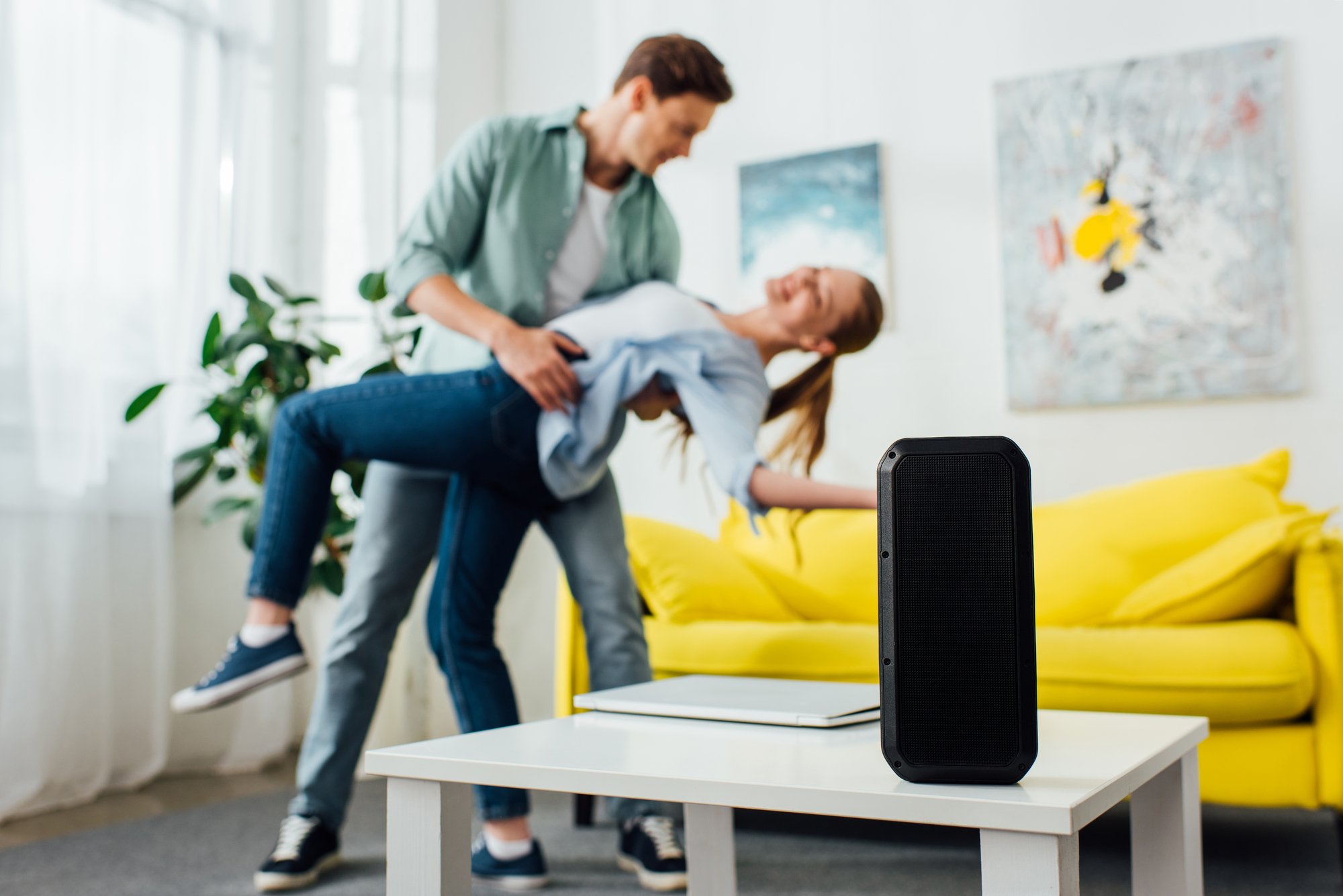 Selective focus of wireless speaker and laptop on coffee table and smiling young couple dancing at home